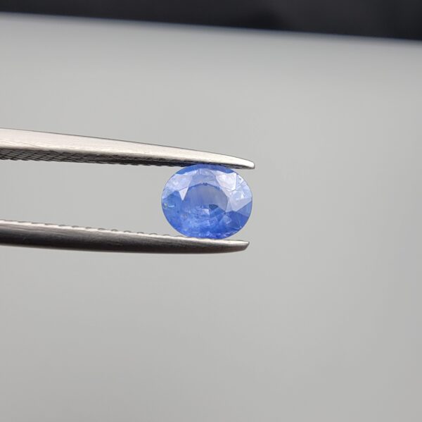 Studio close-up photographs showing the top table face and reverse facet profile of a 0.96 carat oval cut natural blue sapphire loose gemstone held in precision tweezers.