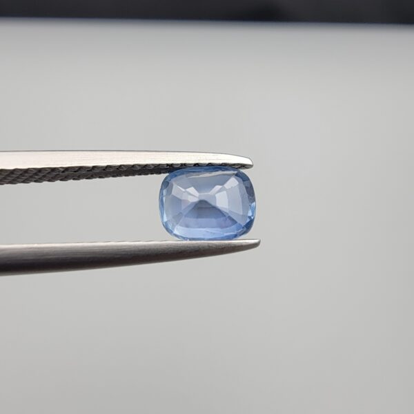 Studio close-up photographs showing the top table face and reverse facet profile of a 1.05 carat cushion cut natural blue sapphire loose gemstone held in precision tweezers.