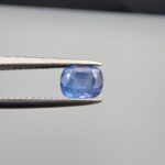 Studio close-up photographs showing the top table face and reverse facet profile of a 1.05 carat cushion cut natural blue sapphire loose gemstone held in precision tweezers.