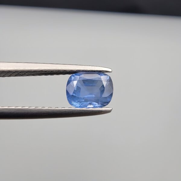 Studio close-up photographs showing the top table face and reverse facet profile of a 1.05 carat cushion cut natural blue sapphire loose gemstone held in precision tweezers.
