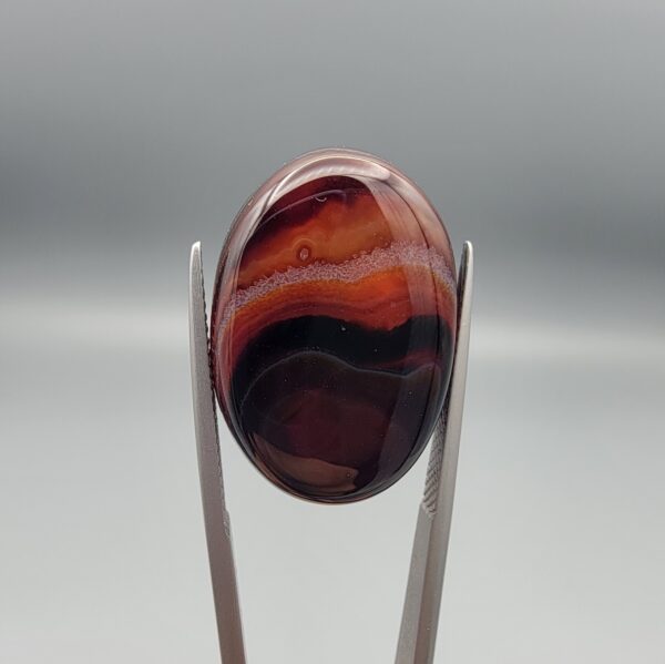 "Natural Carnelian Banded Agate Sulemani Aqeeq gemstone 25.08 carat oval cabochon from Iran held in tweezers showing brown, black, and white natural bands"