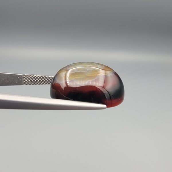 Studio close-up photographs showing the high-domed top surface, side profile, and reverse pattern views of a 17.43 carat oval cabochon natural multi-color agate loose gemstone held in precision tweezers.