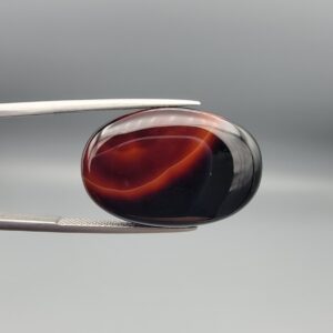 Studio close-up photographs showing the high-domed top surface, contrasting natural color bands, and reverse pattern views of a 25.48 carat oval cabochon natural banded agate loose gemstone held in precision tweezers.