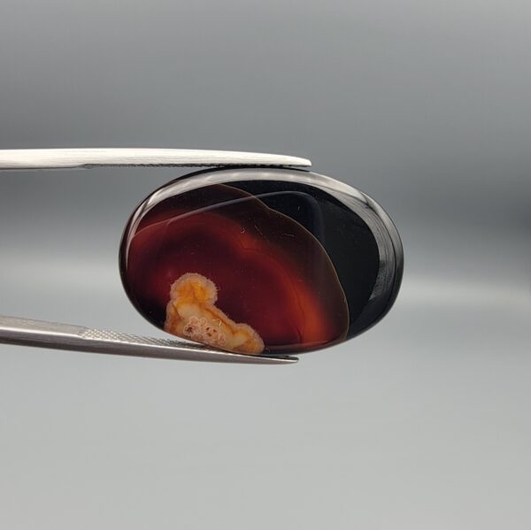 Studio close-up photographs showing the high-domed top surface, contrasting natural color bands, and reverse pattern views of a 25.48 carat oval cabochon natural banded agate loose gemstone held in precision tweezers.