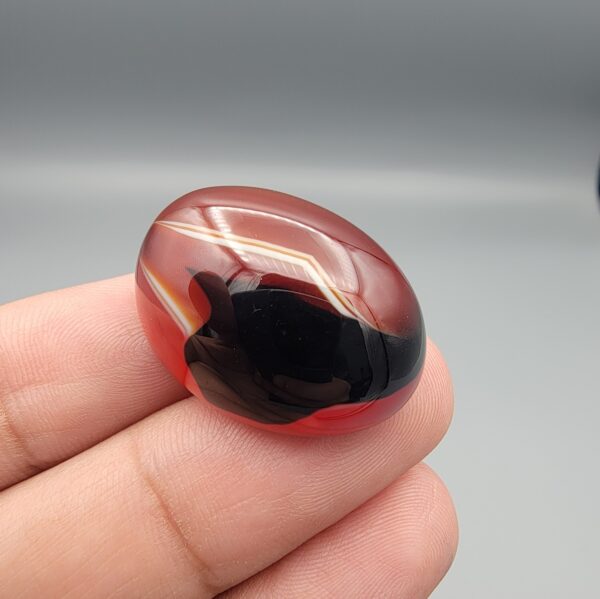 Studio close-up photographs showing the high-domed top surface, contrasting natural color bands, and finger-scale perspective views of a 60.66 carat oval cabochon natural Sulemani agate loose gemstone.