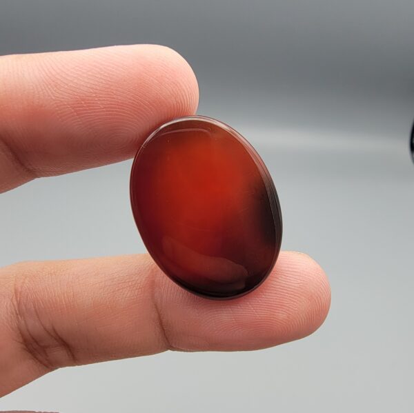 Studio close-up photographs showing the high-domed top surface, contrasting natural color bands, and finger-scale perspective views of a 60.66 carat oval cabochon natural Sulemani agate loose gemstone.