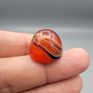 Studio close-up photographs showing the high-domed top surface, distinct natural multi-color banding, and finger-scale perspective views of a 34.28 carat dome-shaped natural carnelian Sulemani agate loose gemstone.