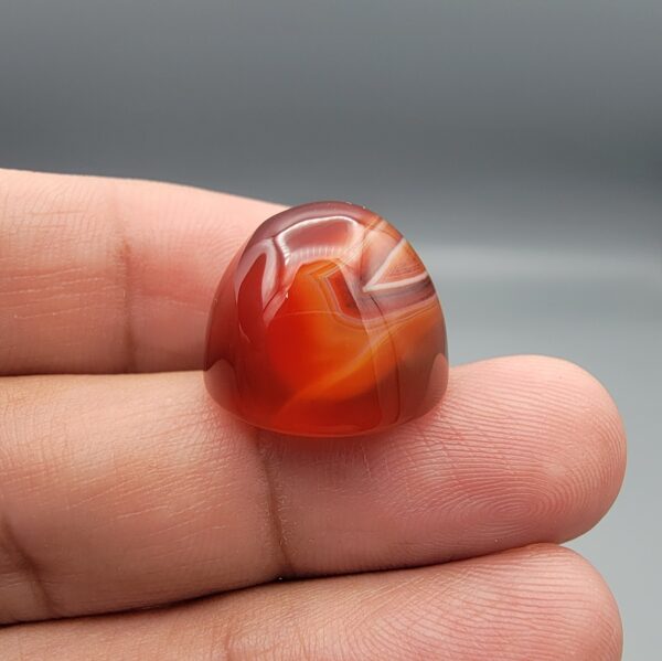 Studio close-up photographs showing the high-domed top surface, distinct natural multi-color banding, and finger-scale perspective views of a 34.28 carat dome-shaped natural carnelian Sulemani agate loose gemstone.