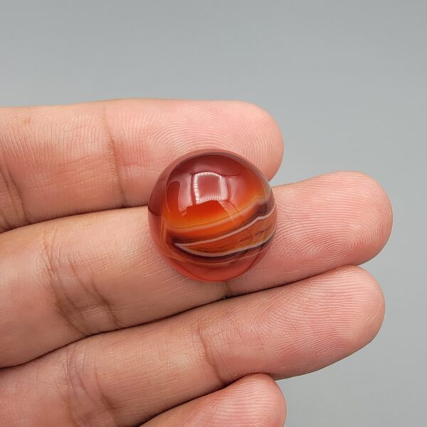 Studio close-up photographs showing the high-domed top surface, distinct natural multi-color banding, and finger-scale perspective views of a 34.28 carat dome-shaped natural carnelian Sulemani agate loose gemstone.