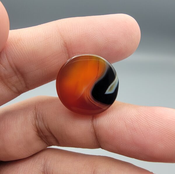 Studio close-up photographs showing the high-domed top surface, distinct natural multi-color banding, and finger-scale perspective views of a 34.28 carat dome-shaped natural carnelian Sulemani agate loose gemstone.