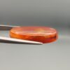 Studio close-up photographs showing the smooth polished dome, side profile featuring distinct internal black banding, and reverse pattern views of a 24.85 carat oval carnelian agate loose gemstone held in precision tweezers.