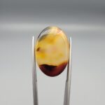 Studio close-up photographs showing the high-domed top surface, contrasting black and orange banding, and orientation views of a 21.68 carat oval cabochon natural chalcedony agate loose gemstone held in precision tweezers.