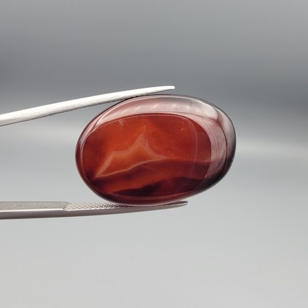 Studio close-up photographs showing the high-domed top surface, deep dark banding, and reverse pattern views of a 28.80 carat oval cabochon natural chalcedony agate loose gemstone held in precision tweezers.