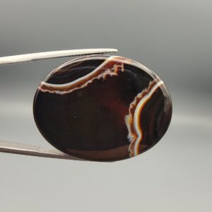 Studio close-up photographs showing the high-domed top surface, deep dark base, intricate organic white and brown banding, and profile thickness views of a 51.94 carat oval cabochon Sulemani Aqeeq agate loose gemstone held in precision tweezers.