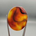 ​Studio close-up photographs showing the high-domed translucent top surface, intricate organic black and white banding formations, and reverse pattern views of a 49.55 carat oval cabochon orangish yellow agate loose gemstone held in precision tweezers.