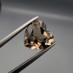 Studio close-up photograph showing the precision facet pattern, premium clarity, and rich brown color of a 3.33 carat heart shape brilliant cut natural smoky quartz loose gemstone held securely in tweezers.
