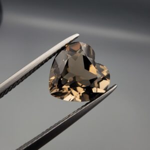 Studio close-up photograph showing the precision facet pattern, premium clarity, and rich brown color of a 3.33 carat heart shape brilliant cut natural smoky quartz loose gemstone held securely in tweezers.