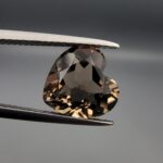 Studio close-up photographs showing the precision facet pattern, premium clarity, and rich brown color of a 3.29 carat heart shape brilliant cut natural smoky quartz loose gemstone held securely in tweezers.