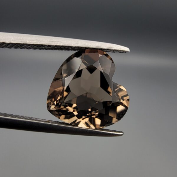 Studio close-up photographs showing the precision facet pattern, premium clarity, and rich brown color of a 3.29 carat heart shape brilliant cut natural smoky quartz loose gemstone held securely in tweezers.