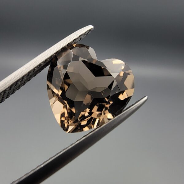 Studio close-up photographs showing the precision facet pattern, premium clarity, and rich brown color of a 3.29 carat heart shape brilliant cut natural smoky quartz loose gemstone held securely in tweezers.