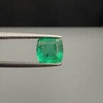 Studio close-up photograph showcasing a cushion-cut 2.12 carat natural green emerald gemstone held securely in precision tweezers, revealing distinctive internal black silk inclusions.