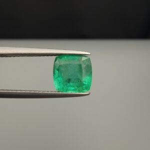 Studio close-up photograph showcasing a cushion-cut 2.12 carat natural green emerald gemstone held securely in precision tweezers, revealing distinctive internal black silk inclusions.