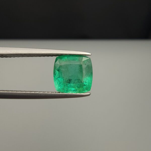 Studio close-up photograph showcasing a cushion-cut 2.12 carat natural green emerald gemstone held securely in precision tweezers, revealing distinctive internal black silk inclusions.
