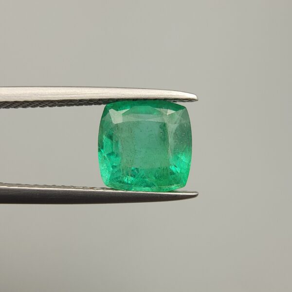 Studio close-up photograph showcasing a cushion-cut 2.12 carat natural green emerald gemstone held securely in precision tweezers, revealing distinctive internal black silk inclusions.