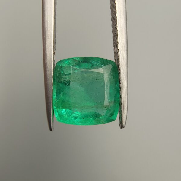 Studio close-up photograph showcasing a cushion-cut 2.12 carat natural green emerald gemstone held securely in precision tweezers, revealing distinctive internal black silk inclusions.