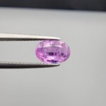 Studio close-up photograph showcasing a 1.55-carat oval-cut natural vivid purple sapphire loose gemstone featuring visible internal feathering and a subtle flash of blue zoning near the lower girdle edge, held securely in precision tweezers.