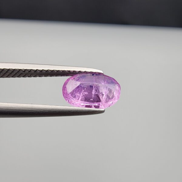 Studio close-up photograph showcasing a 1.55-carat oval-cut natural vivid purple sapphire loose gemstone featuring visible internal feathering and a subtle flash of blue zoning near the lower girdle edge, held securely in precision tweezers.