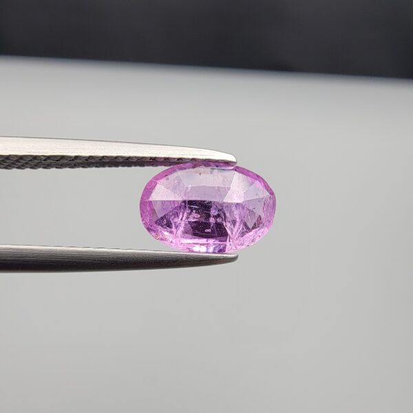 Studio close-up photograph showcasing a 1.55-carat oval-cut natural vivid purple sapphire loose gemstone featuring visible internal feathering and a subtle flash of blue zoning near the lower girdle edge, held securely in precision tweezers.