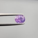 Studio close-up photographs showcasing a 1.44-carat oval-cut natural vivid purple sapphire loose gemstone with visible internal feathering and distinct color zoning, held securely in precision tweezers against a neutral grey background.