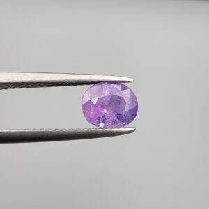 Studio close-up photographs showcasing a 1.44-carat oval-cut natural vivid purple sapphire loose gemstone with visible internal feathering and distinct color zoning, held securely in precision tweezers against a neutral grey background.