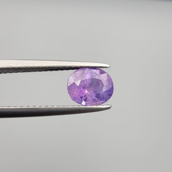 Studio close-up photographs showcasing a 1.44-carat oval-cut natural vivid purple sapphire loose gemstone with visible internal feathering and distinct color zoning, held securely in precision tweezers against a neutral grey background.