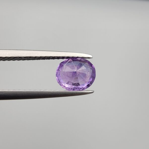 Studio close-up photographs showcasing a 1.44-carat oval-cut natural vivid purple sapphire loose gemstone with visible internal feathering and distinct color zoning, held securely in precision tweezers against a neutral grey background.