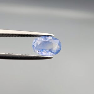 Studio close-up photograph showcasing a 1.38 carat oval-cut natural blue sapphire Neelam loose gemstone held securely in precision tweezers against a neutral grey background.