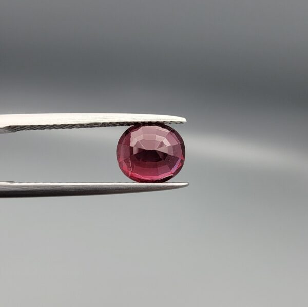 Studio close-up photographs displaying a 3.13-carat oval-cut natural rhodolite garnet gemstone featuring a unique surface inclusion detail on the front, held securely in precision tweezers against a neutral grey background.