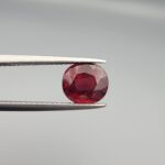 Studio close-up photographs displaying a 3.13-carat oval-cut natural rhodolite garnet gemstone featuring a unique surface inclusion detail on the front, held securely in precision tweezers against a neutral grey background.