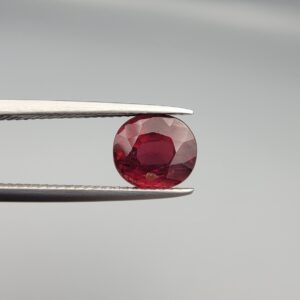 Studio close-up photographs displaying a 3.13-carat oval-cut natural rhodolite garnet gemstone featuring a unique surface inclusion detail on the front, held securely in precision tweezers against a neutral grey background.