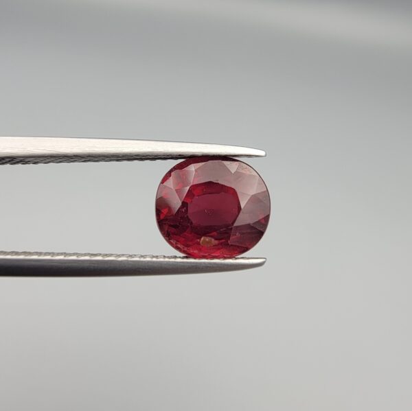 Studio close-up photographs displaying a 3.13-carat oval-cut natural rhodolite garnet gemstone featuring a unique surface inclusion detail on the front, held securely in precision tweezers against a neutral grey background.