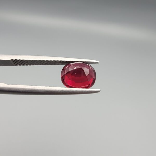 Studio close-up photographs displaying a 2.72-carat oval-cut natural rhodolite garnet gemstone held securely in precision tweezers against a neutral grey background.