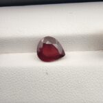 Studio close-up photograph displaying a 2.81-carat pear-cut natural rhodolite garnet gemstone resting on a white textured display surface.