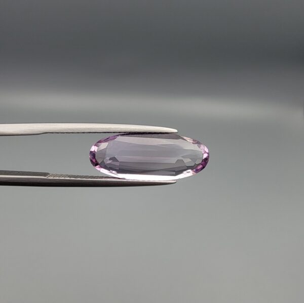 ​Studio close-up photographs displaying a 4.38-carat elongated oval-cut natural amethyst Katela gemstone held securely in precision tweezers against a neutral grey background.