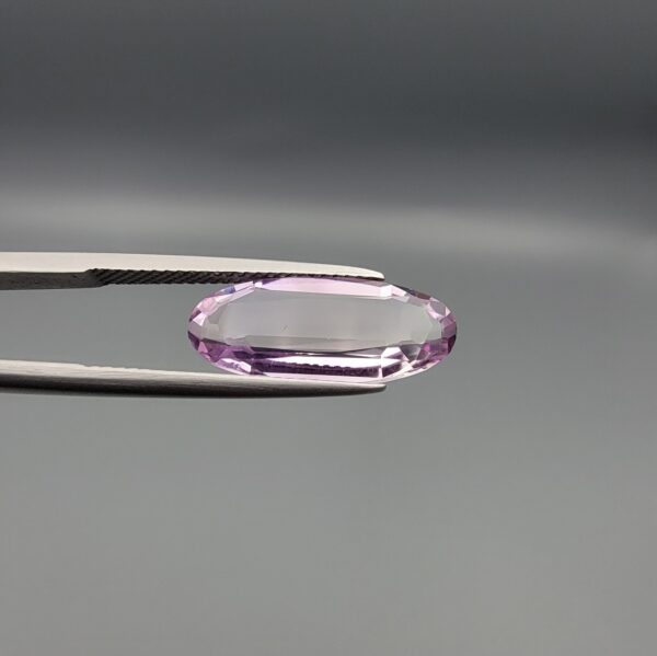 ​Studio close-up photographs displaying a 4.38-carat elongated oval-cut natural amethyst Katela gemstone held securely in precision tweezers against a neutral grey background.