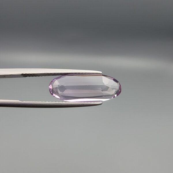 ​Studio close-up photographs displaying a 4.38-carat elongated oval-cut natural amethyst Katela gemstone held securely in precision tweezers against a neutral grey background.