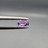 Studio close-up photograph displaying a 0.37-carat oval-cut natural amethyst Katela gemstone held securely in precision tweezers against a neutral grey background.