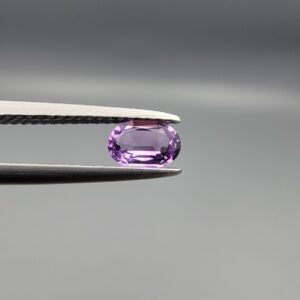 Studio close-up photograph displaying a 0.37-carat oval-cut natural amethyst Katela gemstone held securely in precision tweezers against a neutral grey background.