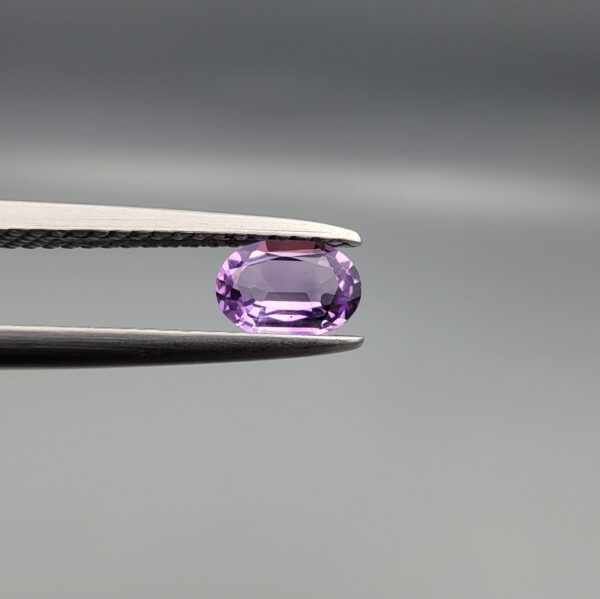 Studio close-up photograph displaying a 0.37-carat oval-cut natural amethyst Katela gemstone held securely in precision tweezers against a neutral grey background.