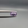 Studio close-up photograph displaying a 0.37-carat oval-cut natural amethyst Katela gemstone held securely in precision tweezers against a neutral grey background.